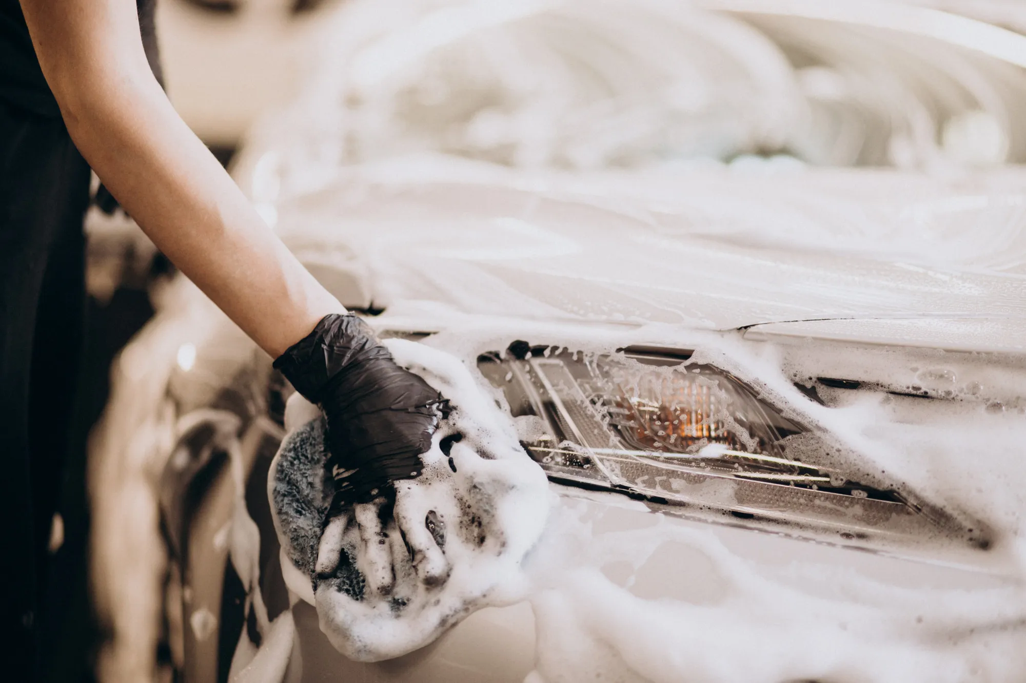 A gloved hand washing a car headlight with a soapy sponge
