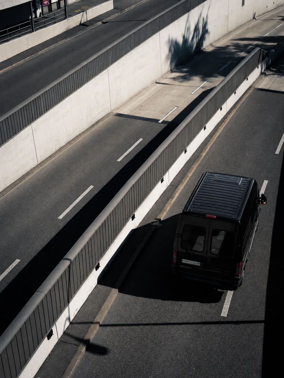 Black van driving in a sunlit urban divided road with trees and shadows.